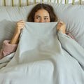 A young woman peeking from behind her Avec bedding linen-blend sheets in the morning